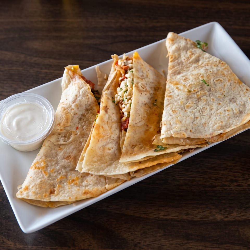 Four quesadilla wedges with chicken and cheese, served with a side of sour cream on a white plate.