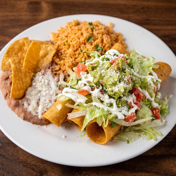 A plate of Mexican food with taquitos, rice, refried beans, chips, guacamole, and sour cream.